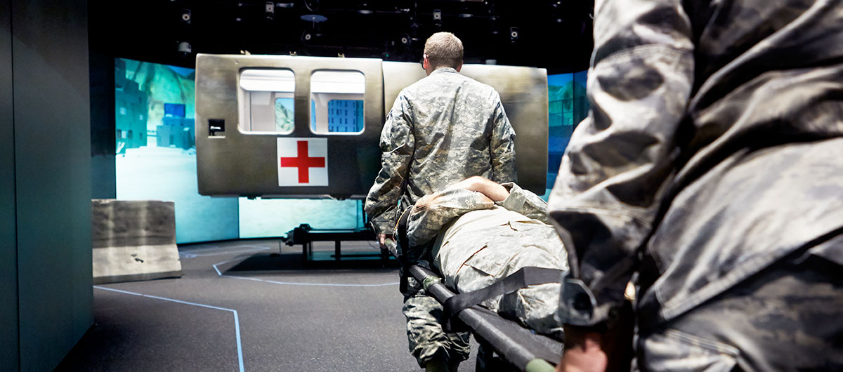 Military medical students carry a patient toward a simulator.