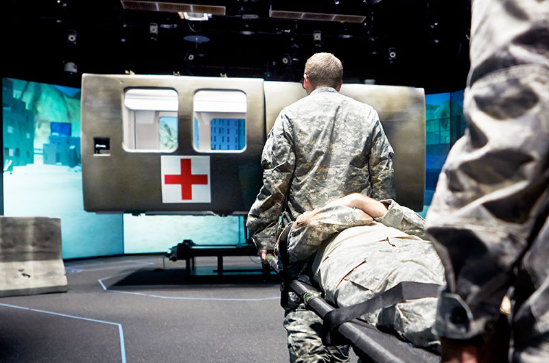 Military medical students carry a patient toward a simulator.
